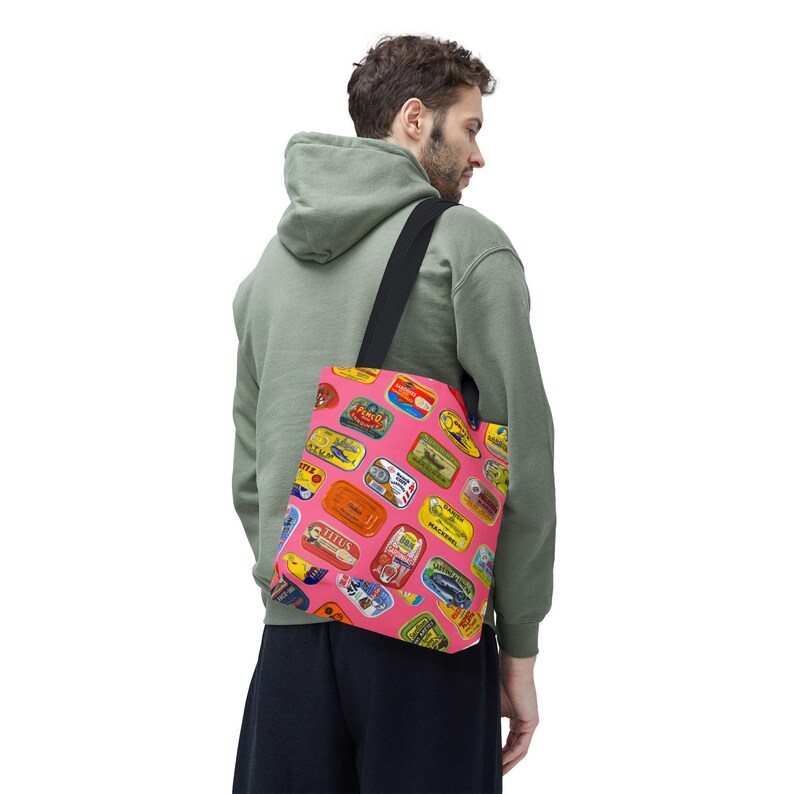 A person wearing a green hoodie and black shorts carries a pink tote bag with sardine can labels over their left shoulder, viewed from behind.