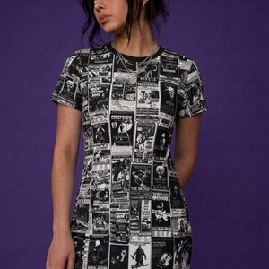 A model from the front wearing a black t-shirt dress with a black-and-white horror poster all-over print, fishnet tights, and dark hair against a purple wall.