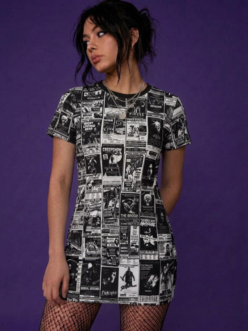 A model from the front wearing a black t-shirt dress with a black-and-white horror poster all-over print, fishnet tights, and dark hair against a purple wall.