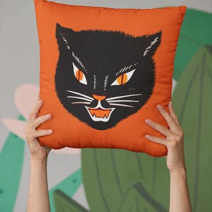 May include: Orange decorative pillow featuring a black cat face with orange eyes and a white mouth. The cat's face is centered on the pillow, which is held up by two hands. The pillow is a Halloween-themed decorative item.