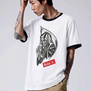 A man posing in a white ringer t-shirt that has black collar and sleeve bands, showcasing a grim reaper graphic and red text reading Killin it.