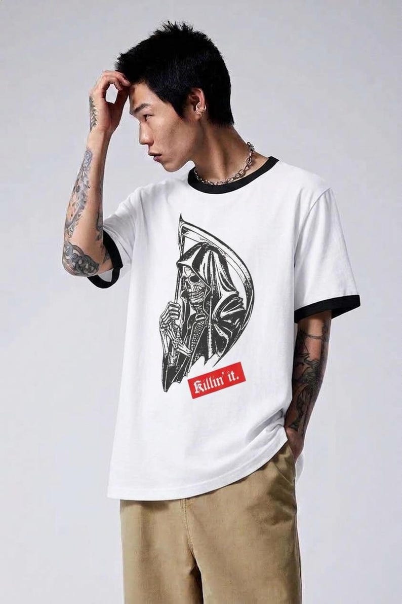 A man posing in a white ringer t-shirt that has black collar and sleeve bands, showcasing a grim reaper graphic and red text reading Killin it.