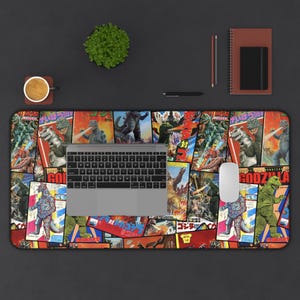 May include: A rectangular desk mat featuring a collage of vintage Godzilla movie posters in various colors. A laptop, mouse, and other office supplies are on a dark surface. The mat's design includes Japanese text and illustrations of Godzilla.