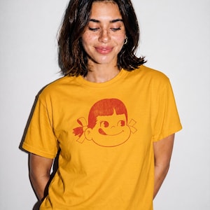 A person with short dark hair wears a mustard yellow t-shirt with a red graphic of a girl's face with pigtails and a winking expression, against a white background.