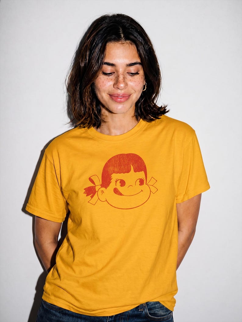 A person with short dark hair wears a mustard yellow t-shirt with a red graphic of a girl's face with pigtails and a winking expression, against a white background.