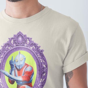 May include: Cream-coloured t-shirt featuring a framed illustration of a superhero figure. The illustration is set against a lime green background within a purple ornate frame. The superhero is depicted in red, blue, and silver, giving the middle finger.
