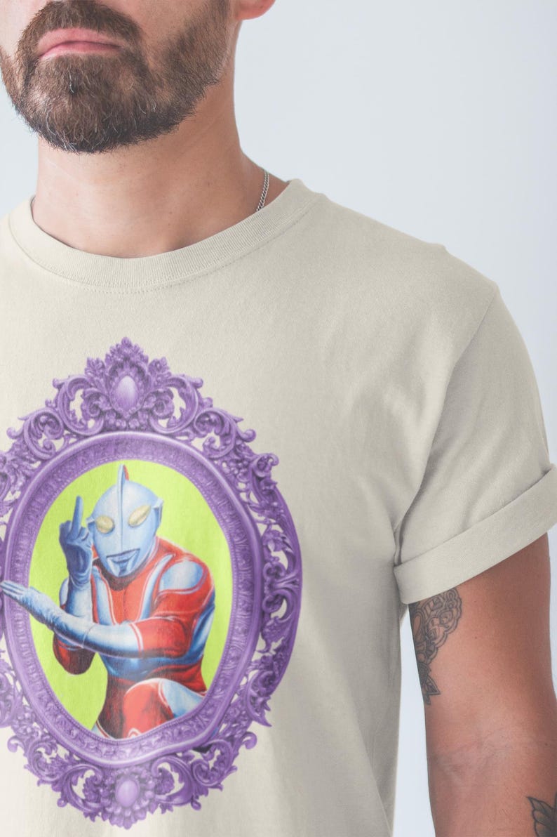 May include: Cream-coloured t-shirt featuring a framed illustration of a superhero figure. The illustration is set against a lime green background within a purple ornate frame. The superhero is depicted in red, blue, and silver, giving the middle finger.
