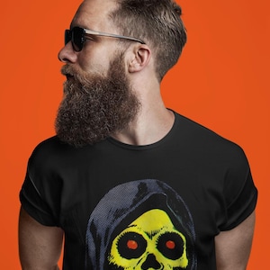 May include: A black t-shirt featuring a yellow skull design with a black hood. The skull has red eyes and a detailed jawline. The person wearing the shirt has a full beard, sunglasses, and is set against an orange background.
