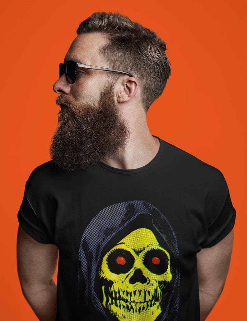 May include: A black t-shirt featuring a yellow skull design with a black hood. The skull has red eyes and a detailed jawline. The person wearing the shirt has a full beard, sunglasses, and is set against an orange background.