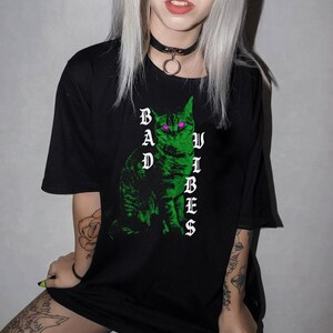 A person with blonde hair and a black choker sits while wearing a black t-shirt printed with a green cat and white text.