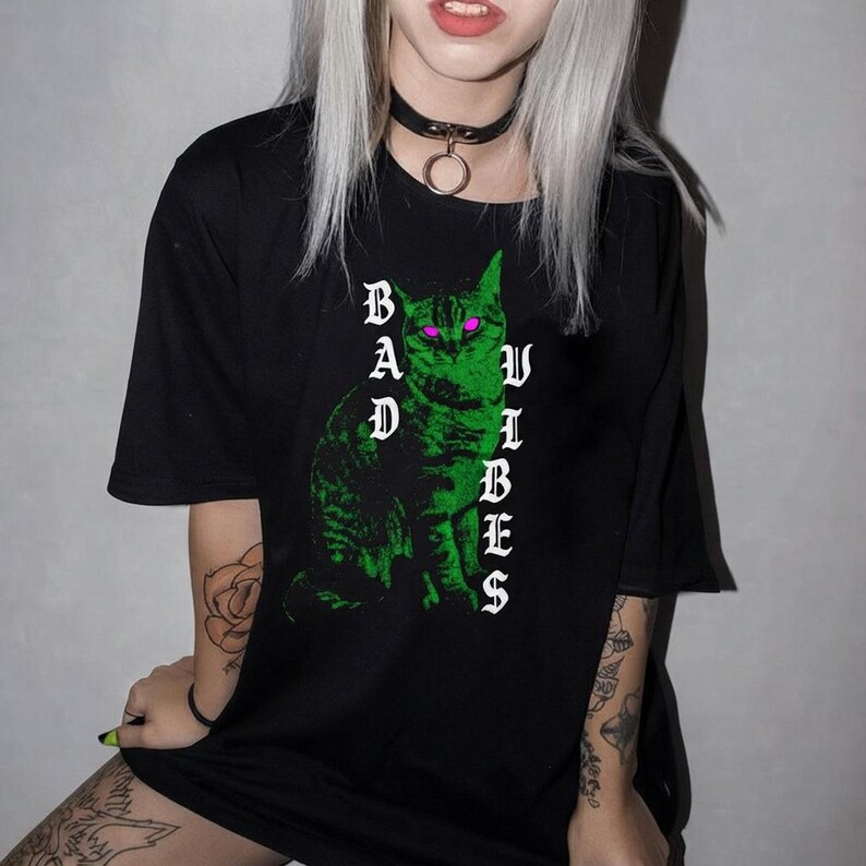 A person with blonde hair and a black choker sits while wearing a black t-shirt printed with a green cat and white text.