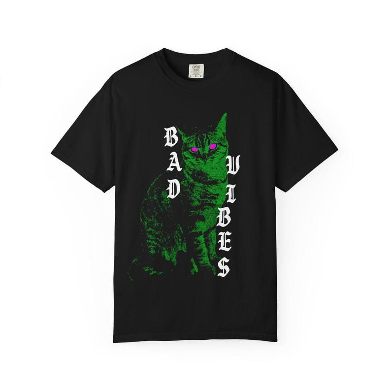 A black t-shirt laid flat, featuring a graphic of a green cat with glowing purple eyes and white gothic text reading Bad Vibes.