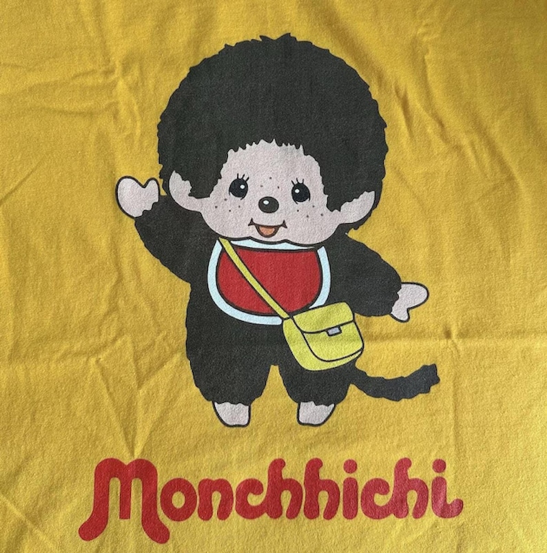 A close-up of a yellow fabric showing a Monchhichi graphic character wearing a red collar and yellow shoulder bag above the text Monchhichi.