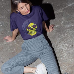 A person with long brown hair sits on the ground wearing a purple t-shirt with a yellow skull in a blue hood and striped pants.