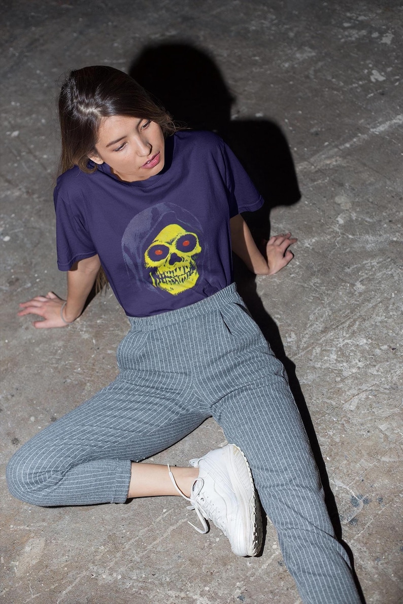 A person with long brown hair sits on the ground wearing a purple t-shirt with a yellow skull in a blue hood and striped pants.