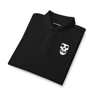 A neatly folded black polo shirt with a white skull embroidery on the left chest, isolated on a white background.