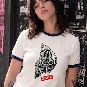 A person leaning against a wall wearing a white ringer t-shirt featuring dark blue trim and a black and white grim reaper illustration with a red Killin it label.