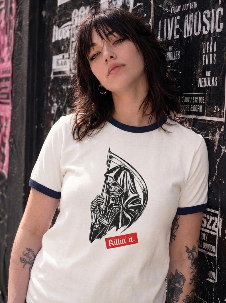 A person leaning against a wall wearing a white ringer t-shirt featuring dark blue trim and a black and white grim reaper illustration with a red Killin it label.