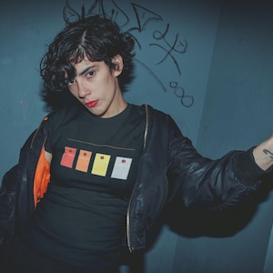Puede incluir: Camiseta negra con un diseño gráfico de cuatro formas rectangulares en rojo, naranja, amarillo y blanco. La persona lleva una chaqueta bomber negra con forro naranja. El fondo es una pared azul con graffiti.