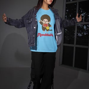 A woman wearing a denim jacket, light blue Monchhichi t-shirt, black cargo pants, and red glasses poses in front of a window.