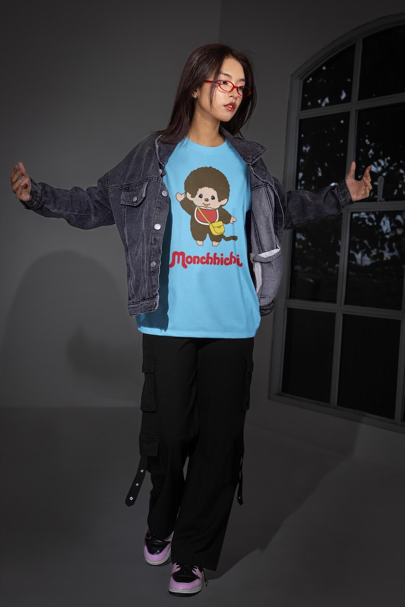 A woman wearing a denim jacket, light blue Monchhichi t-shirt, black cargo pants, and red glasses poses in front of a window.