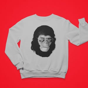 K&ouml;nnte beinhalten: Wei&szlig;es Sweatshirt mit einem schwarz-wei&szlig;en Aufdruck eines Affen mit Sonnenbrille. Das Affenmotiv befindet sich auf der Vorderseite des lang&auml;rmeligen Sweatshirts. Der Hintergrund ist einfarbig rot.