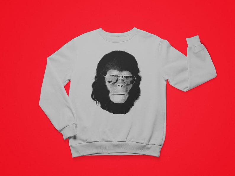 K&ouml;nnte beinhalten: Wei&szlig;es Sweatshirt mit einem schwarz-wei&szlig;en Aufdruck eines Affen mit Sonnenbrille. Das Affenmotiv befindet sich auf der Vorderseite des lang&auml;rmeligen Sweatshirts. Der Hintergrund ist einfarbig rot.