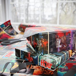 May include: Two wrapped gift boxes with black elastic bands, featuring horror movie-themed wrapping paper. The paper displays movie posters with titles like "C.H.U.D." and "House." A pair of scissors and a roll of matching paper are also visible.