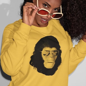 K&ouml;nnte beinhalten: Senfgelbes Sweatshirt mit einem schwarz-wei&szlig;en Aufdruck eines Affen mit Sonnenbrille. Die Person tr&auml;gt eine Sonnenbrille mit roten Gl&auml;sern. Das Sweatshirt ist ein l&auml;ssiges Langarm-Kleidungsst&uuml;ck.