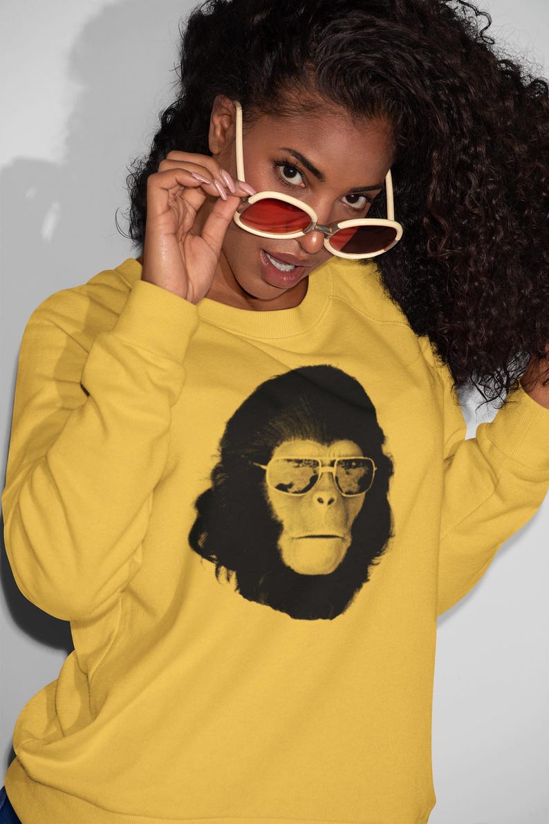 K&ouml;nnte beinhalten: Senfgelbes Sweatshirt mit einem schwarz-wei&szlig;en Aufdruck eines Affen mit Sonnenbrille. Die Person tr&auml;gt eine Sonnenbrille mit roten Gl&auml;sern. Das Sweatshirt ist ein l&auml;ssiges Langarm-Kleidungsst&uuml;ck.