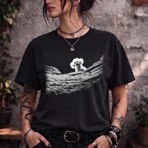 May include: A black short-sleeved t-shirt featuring a graphic print of a small white fluffy dog sitting on a textured, weathered log. The shirt has a crew neckline and a relaxed fit, showcasing a detailed monochromatic illustration of the canine.