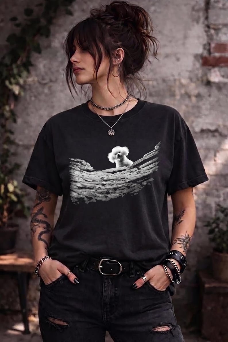 May include: A black short-sleeved t-shirt featuring a graphic print of a small white fluffy dog sitting on a textured, weathered log. The shirt has a crew neckline and a relaxed fit, showcasing a detailed monochromatic illustration of the canine.