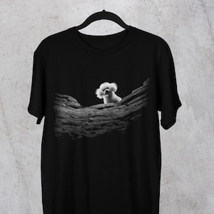 May include: A black short-sleeved t-shirt featuring a central graphic print. The design displays a small, fluffy white dog peeking over a textured, dark grey rock formation. The shirt has a classic crew neckline and is displayed on a black hanger.