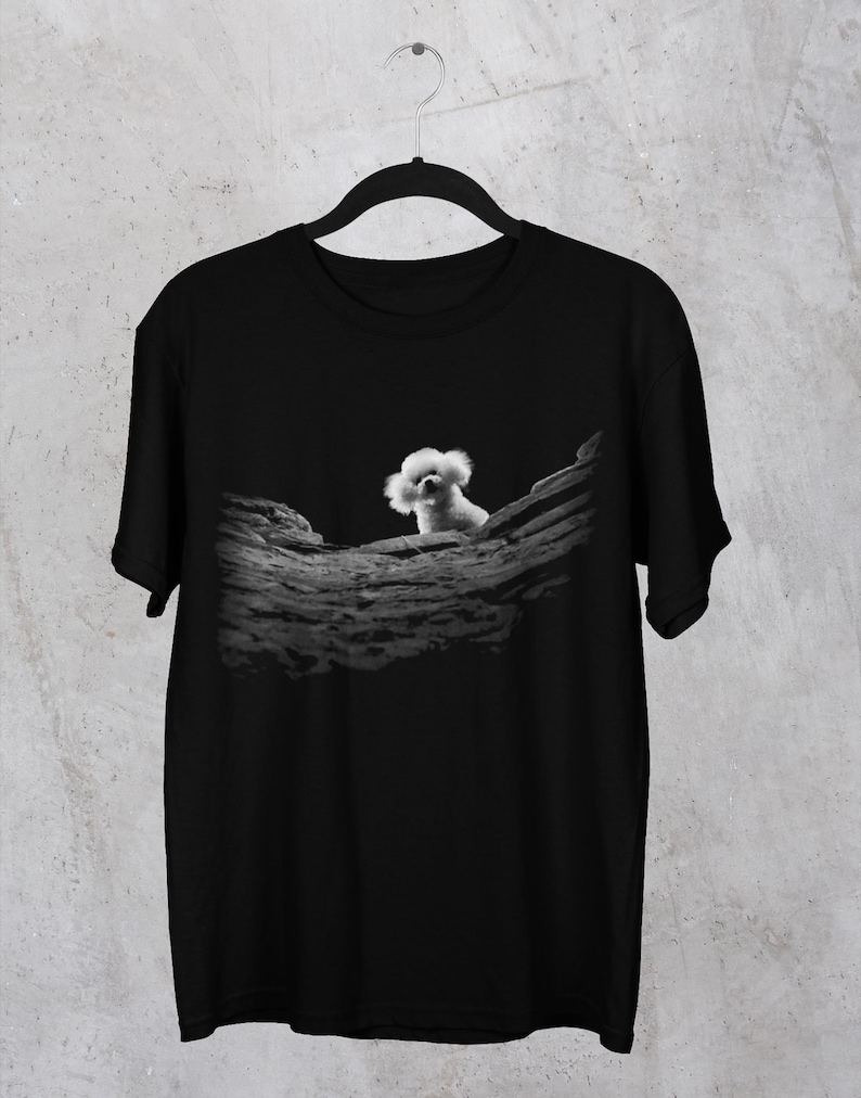 May include: A black short-sleeved t-shirt featuring a central graphic print. The design displays a small, fluffy white dog peeking over a textured, dark grey rock formation. The shirt has a classic crew neckline and is displayed on a black hanger.