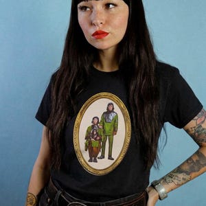 May include: A black short-sleeve t-shirt featuring a central oval graphic print of three figures in green and brown outfits enclosed in an ornate gold frame. The design depicts two standing individuals and one seated figure, all styled in a retro aesthetic.