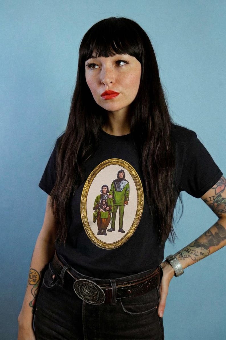 May include: A black short-sleeve t-shirt featuring a central oval graphic print of three figures in green and brown outfits enclosed in an ornate gold frame. The design depicts two standing individuals and one seated figure, all styled in a retro aesthetic.