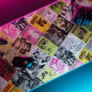 May include: A rectangular gaming mouse pad with a collage of vintage concert posters in various colors. A black mouse, a game controller, and headphones are on the pad. The pad has a neon light border.