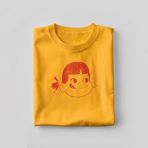 A single folded mustard yellow t-shirt with a red graphic of a cartoon girl's face with a bow in her hair, looking to the side.