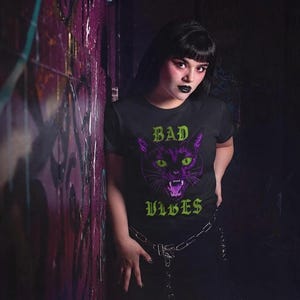 A person with dark hair wearing the black t-shirt featuring the purple cat and green text leaning against a dark brick wall.