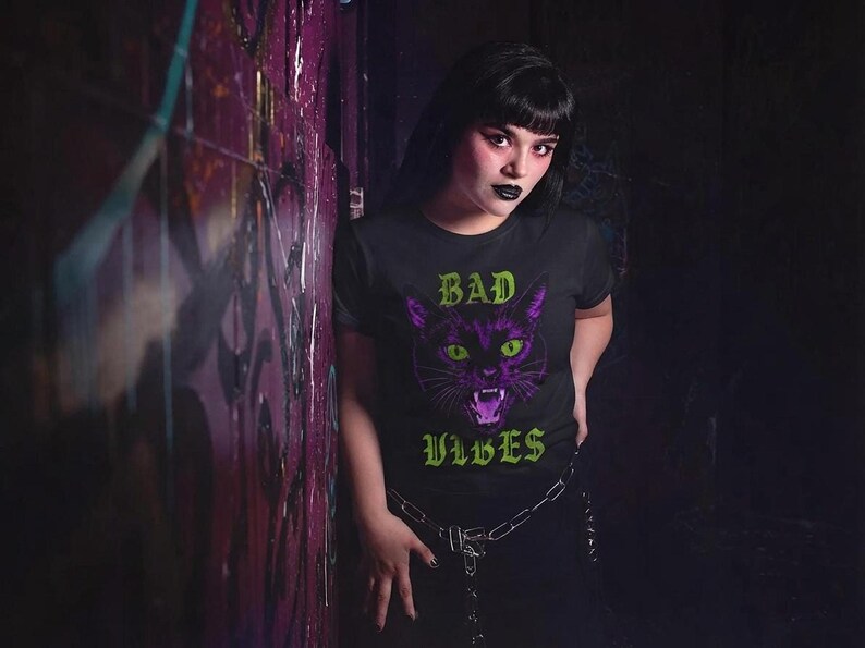 A person with dark hair wearing the black t-shirt featuring the purple cat and green text leaning against a dark brick wall.