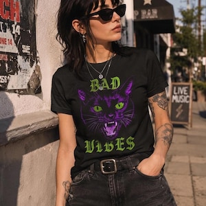 A person wearing sunglasses and the black t-shirt with the graphic cat design poses on a sunny sidewalk next to a building.