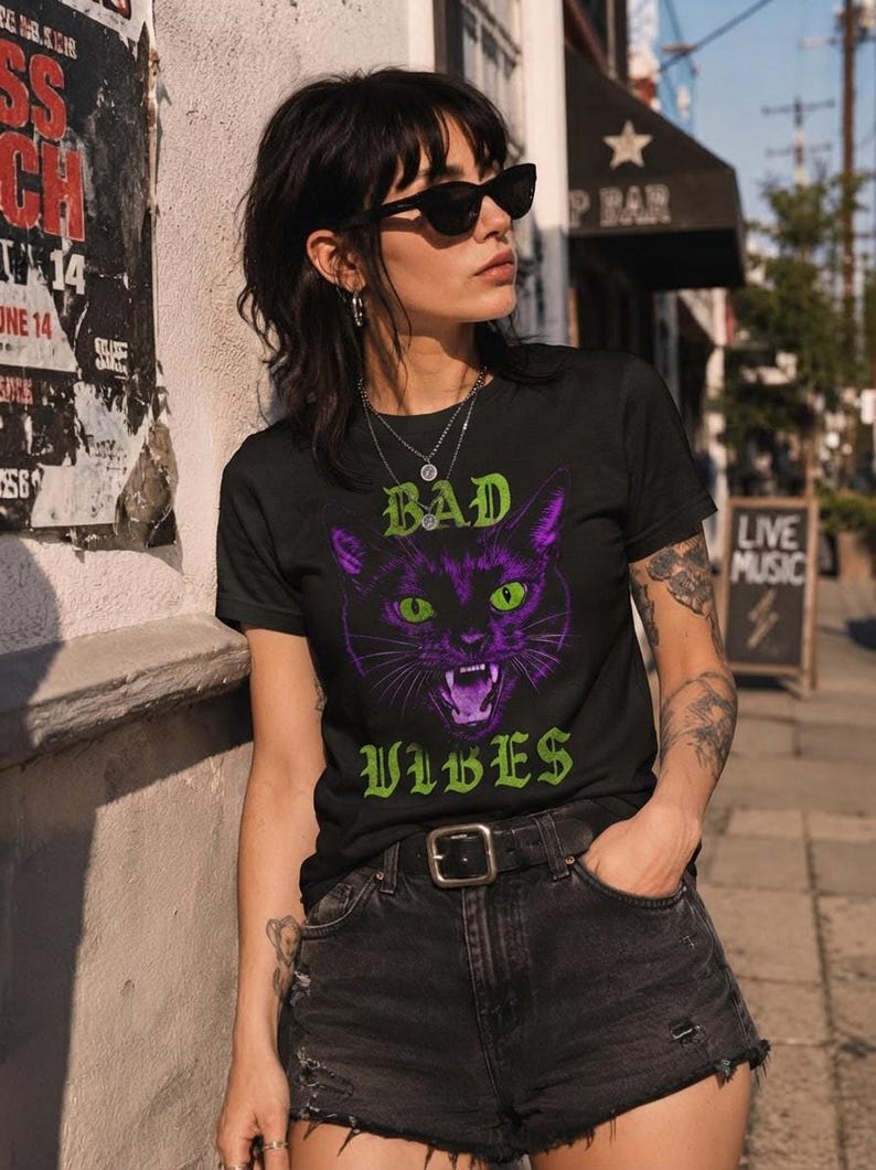 A person wearing sunglasses and the black t-shirt with the graphic cat design poses on a sunny sidewalk next to a building.