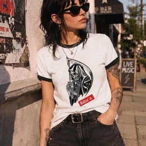 A person wearing sunglasses and a white ringer tee with black trim, printed with a black grim reaper graphic and a red text box that reads Killin it.