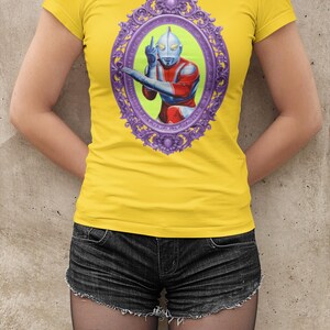 May include: Yellow t-shirt with a framed graphic of a superhero in red, blue, and silver. The oval frame is purple. The shirt is worn with black denim shorts.