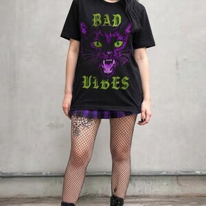 A model standing against a concrete wall wearing the black t-shirt, a purple plaid skirt, fishnet stockings, and platform boots.