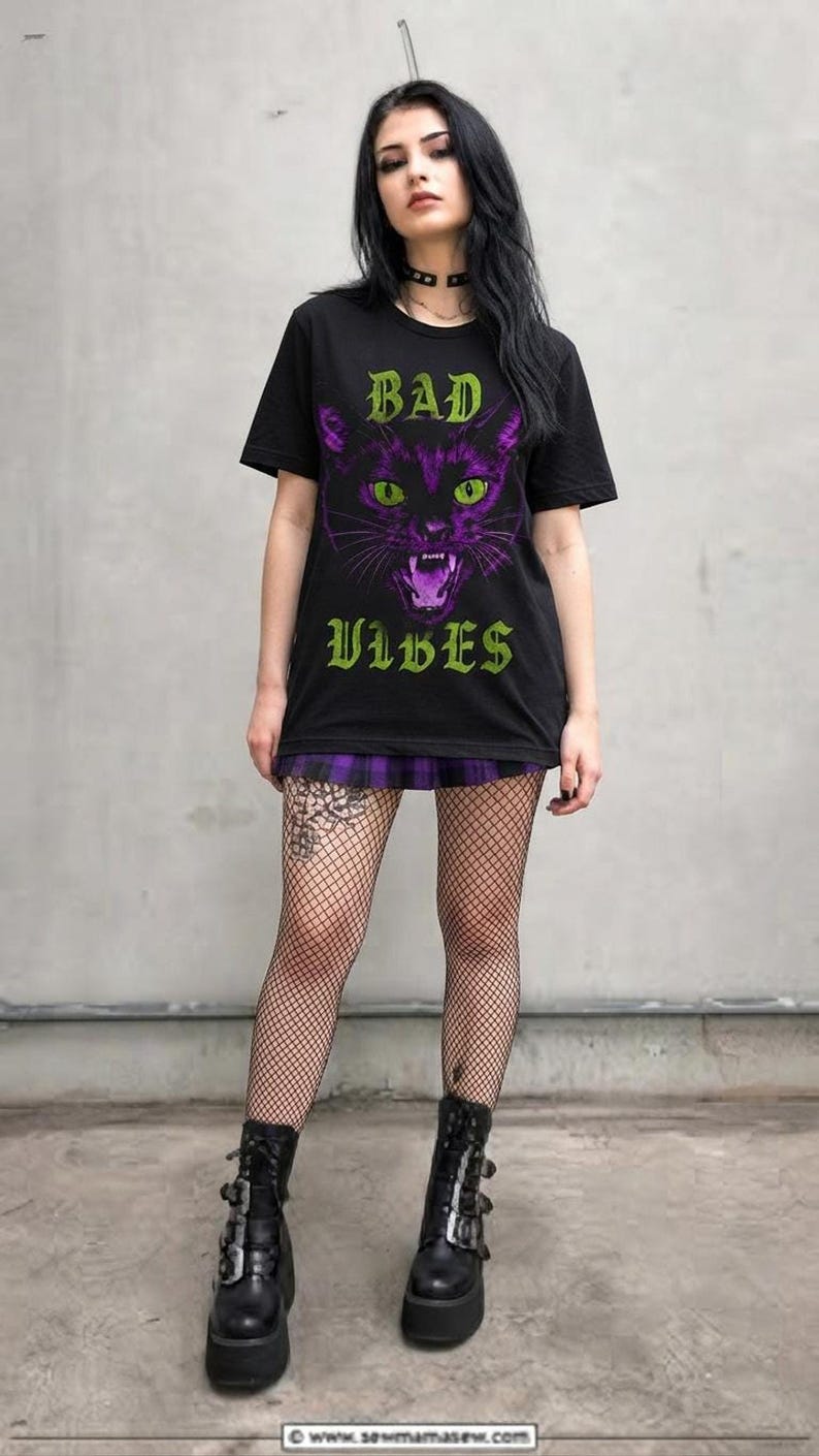 A model standing against a concrete wall wearing the black t-shirt, a purple plaid skirt, fishnet stockings, and platform boots.
