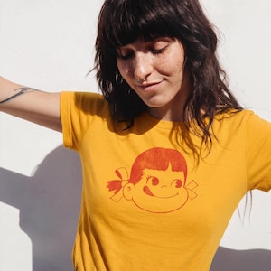 A person with dark hair and a nose ring wears a mustard yellow t-shirt with a red graphic of a girl's face with pigtails and a winking expression, paired with a blue patterned skirt, against a white wall.