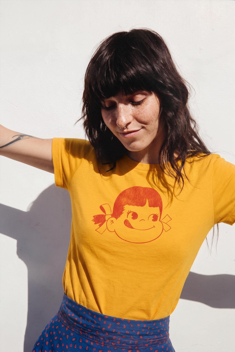 A person with dark hair and a nose ring wears a mustard yellow t-shirt with a red graphic of a girl's face with pigtails and a winking expression, paired with a blue patterned skirt, against a white wall.