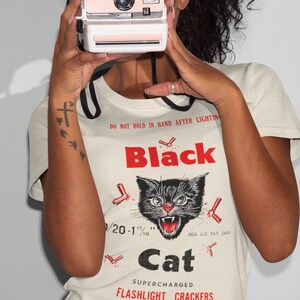 May include: A cream-coloured t-shirt with a graphic of a black cat and the text "Black Cat Flashlight Crackers." The shirt also displays the text "Made in Macau."