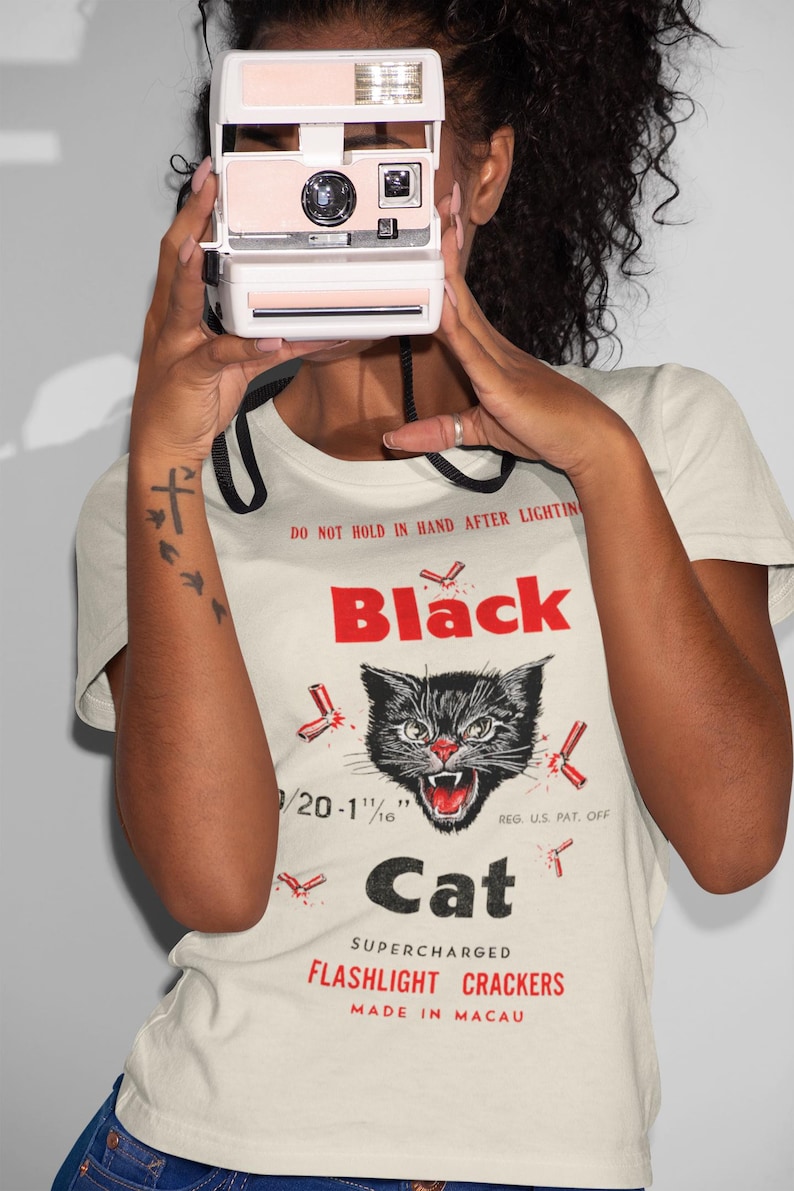 May include: A cream-coloured t-shirt with a graphic of a black cat and the text "Black Cat Flashlight Crackers." The shirt also displays the text "Made in Macau."